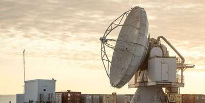 A Base Espacial Pituffik (antiga Base A&eacute;rea de Thule) &eacute; mostrada no norte da Groenl&acirc;ndia, em 4 de outubro de 2023. A imagem mostra uma grande antena parab&oacute;lica em frente a cont&ecirc;ineres, com um c&eacute;u alaranjado e nublado ao fundo. Foto: Thomas Traasdahl/Ritzau Scanpix/AFP via Getty Images / BBC News Brasil