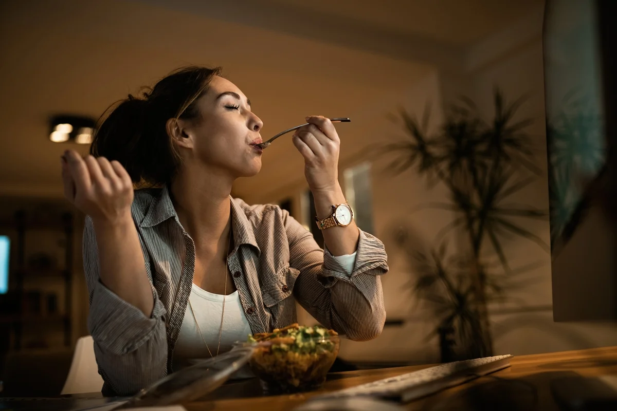Vista de baixo para cima de uma jovem mulher comendo salada com os olhos fechados enquanto usa o computador à noite em casa. Metrópoles