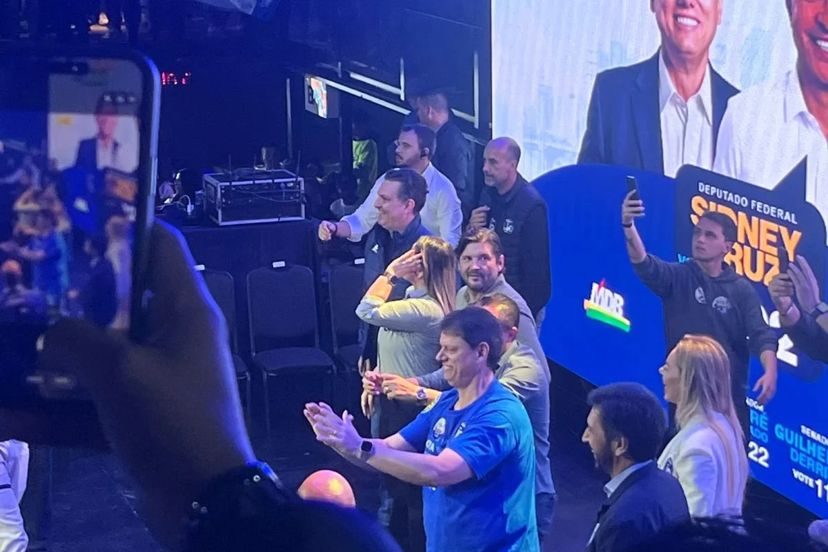 Imagens coloridas mostram o governador Tarcísio de Freitas batendo palmas sobre palco durante evento de campanha