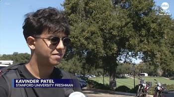 Un joven con gafas de sol oscuras y camiseta negra habla frente a un micrófono en un entorno exterior con árboles