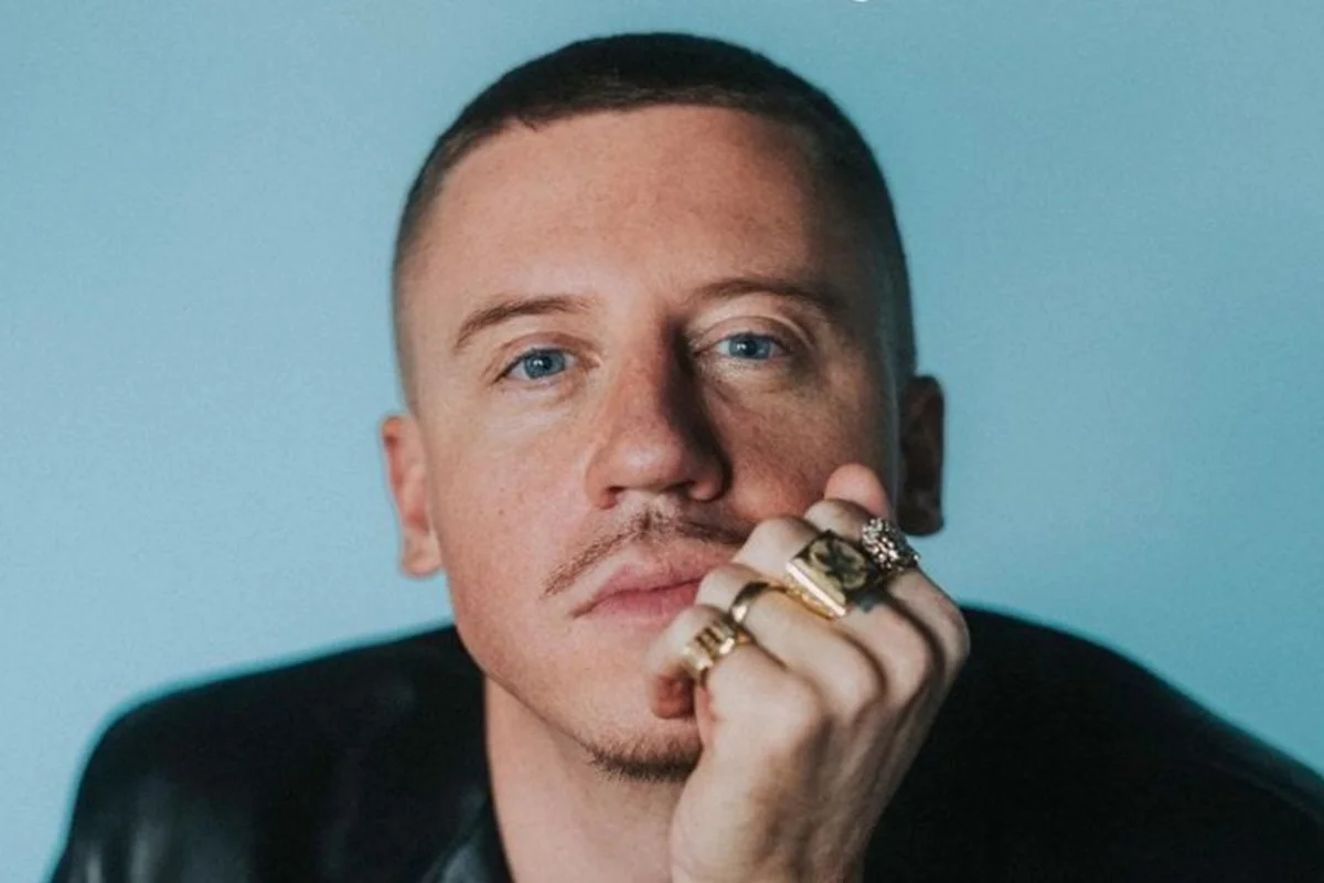 Macklemore em foto colorida. Ele é um homem branco, com olhos azuis, bigode fino e cabelo curto. O fundo da foto é azul - Metrópoles