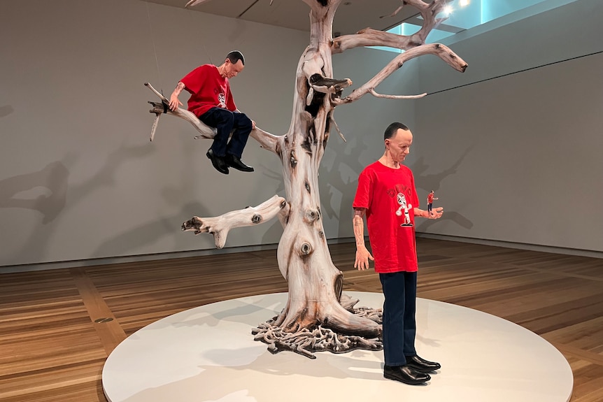 A sculpture of a man holding a tiny of himself in his palm, with a full-sized version of him in a tree behind him.