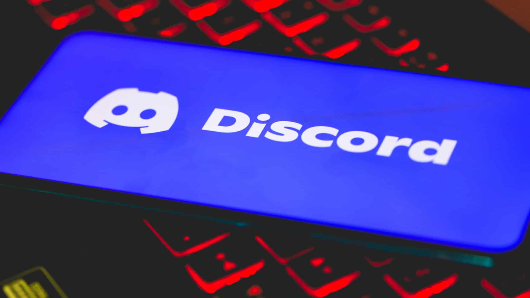 Logo do Discord na tela de um smartphone na diagonal que esta em cima do teclado iluminado de um computador