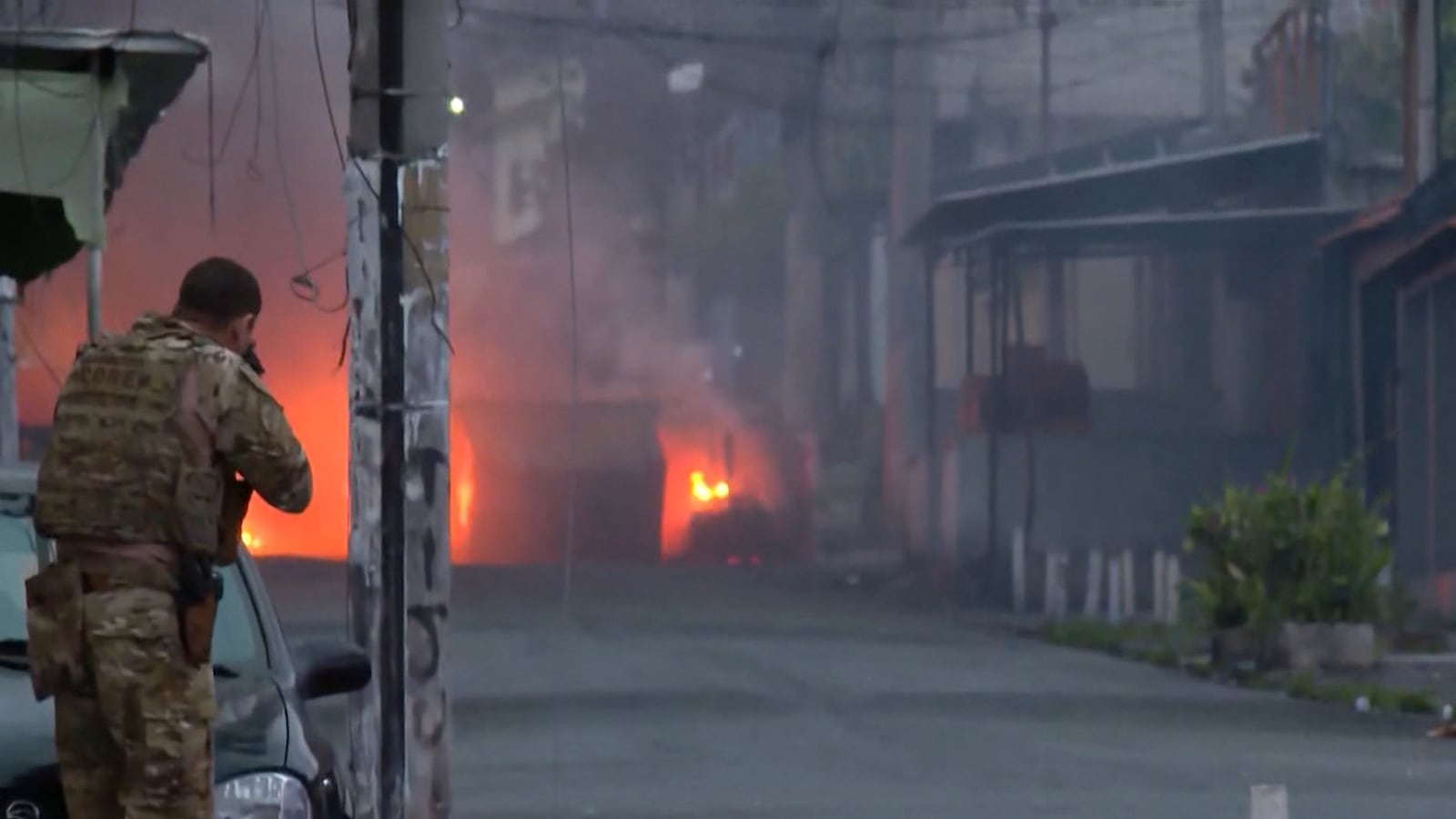 Pessoa em uniforme camuflado se protege atrás de um carro enquanto aponta uma arma. Ao fundo, fogo intenso no meio da rua gera fumaça espessa que encobre parte da cena. O ambiente urbano mostra prédios e fios elétricos cruzando o espaço.
