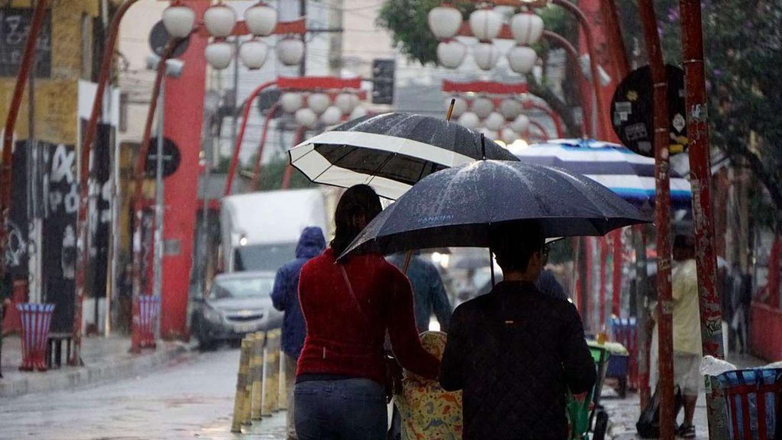 Uma fotografia retangular, tirada ao n&iacute;vel dos olhos, mostrando uma rua movimentada em um dia chuvoso no bairro da Liberdade, em S&atilde;o Paulo. &Agrave; esquerda, uma mulher veste uma blusa de manga comprida vermelha e cal&ccedil;a jeans escura. Ela segura um guarda-chuva aberto de cor preta por fora e branca por dentro. &Agrave; direita, um homem segura um grande guarda-chuva totalmente preto, inclinado sobre a sua cabe&ccedil;a.