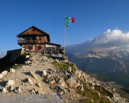 A wooden hut on a peak, with an Italian flag flying and other mountains in the background