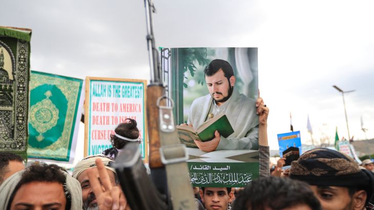 A dense crowd of men fills a street, many raising rifles above their heads, while one holds up a large printed portrait of a bearded man in a turban.
