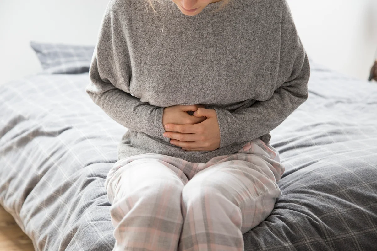 Mulher doente, vestida com roupa de casa cinza, sentada na cama, com as mãos sobre o estômago, sofrendo de dor intensa. Conceito de dor de estômago, cólica ou intoxicação alimentar. Metrópoles